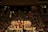 Several sumo wrestlers are gathered in a circle on a sumo wrestling ring surrounded by a large crowd of spectators in a dimly lit arena. The wrestlers are wearing traditional mawashi belts of various colors, and the scene is lit from above, creating a focused and intense atmosphere.