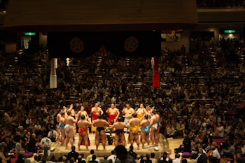 Several sumo wrestlers are gathered in a circle on a sumo wrestling ring surrounded by a large crowd of spectators in a dimly lit arena. The wrestlers are wearing traditional mawashi belts of various colors, and the scene is lit from above, creating a focused and intense atmosphere.