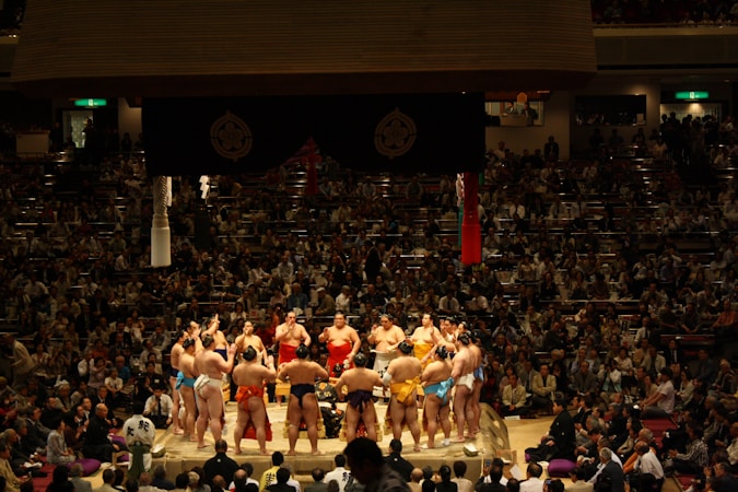 Several sumo wrestlers are gathered in a circle on a sumo wrestling ring surrounded by a large crowd of spectators in a dimly lit arena. The wrestlers are wearing traditional mawashi belts of various colors, and the scene is lit from above, creating a focused and intense atmosphere.