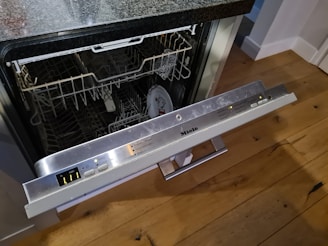 An open dishwasher with visible control buttons on a metallic panel. The interior contains empty racks and a partially visible plate inside. The dishwasher is built into a wooden kitchen with a granite countertop.