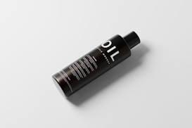A sleek black bottle with the word 'OIL' prominently displayed in large white letters. The bottle has a simple, modern design with additional text details in smaller white font. It sits on a light, neutral background and casts a soft shadow, giving it a floating effect.