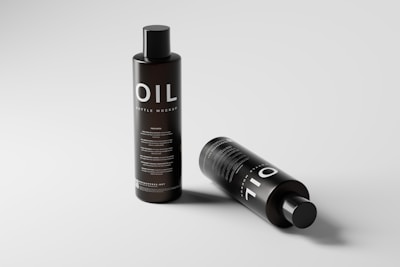 Two black cylindrical bottles are displayed against a light gray background. One bottle stands upright while the other is lying horizontally. The label on each bottle reads 'OIL' in large white letters followed by smaller text that reads 'BOTTLE MOCKUP'. The overall design is sleek and minimalistic.