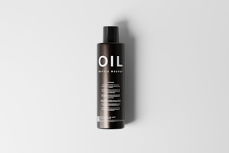 A tall, black cylindrical bottle with a sleek design is placed upright on a light gray background. The bottle features the word 'OIL' prominently displayed in large, white letters at the top, followed by the smaller text 'BOTTLE MOCKUP'. There are multiple lines of text below, which appear to provide additional details, likely about the product or its usage.