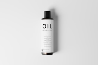 A cylindrical bottle mockup with a black cap and a minimalistic label displaying the word 'OIL' in bold letters. The label features additional text in smaller font, and a QR code is visible at the bottom. The bottle is presented against a plain, light gray background.