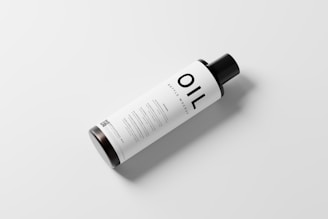 A bottle with a minimalist design labeled 'OIL BOTTLE MOCKUP' rests on a plain white surface. The bottle is cylindrical with a black cap and white label featuring black text and a QR code.