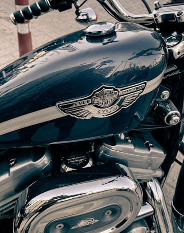 A close-up view of a motorcycle's fuel tank and engine. The fuel tank prominently displays the Harley-Davidson emblem and logo, with a metallic finish. The engine parts are visible beneath, featuring chrome and metal components, reflecting light and surroundings.