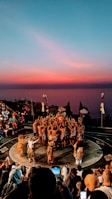 A group of people arranged in a circle are performing a traditional dance outdoors at sunset. The sky has hues of pink, orange, and blue, setting a dramatic backdrop. The dancers are wearing traditional attire, including checked sarongs, and some carry props or wear headdresses. Spectators are seated around the circular performance area, some taking photos or videos. The landscape includes silhouetted structures and natural elements, adding to the cultural ambiance.