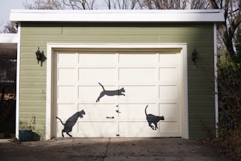 A garage with a light green exterior and a cream-colored, segmented garage door. Three black cat silhouettes are painted on the door, depicting different poses of cats in motion. Small black wall-mounted lanterns are on either side of the door, and bare trees are visible in the background.