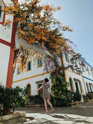 A person in a patterned dress is walking through a picturesque courtyard filled with colorful flowers and lush green plants. The building in the background has a Mediterranean architectural style with geometric designs and arches, adorned with vibrant orange and purple bougainvillea.