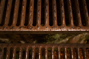 A close-up view of a rusted, metal grate with parallel slats that have circular perforations along the edges. The surface shows signs of weathering and oxidation, giving it a gritty appearance.