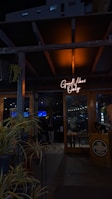 A dimly lit entrance to a sports bar with a glowing neon sign reading 'Good Vibes Only' above the door. The interior appears to be busy, with people visible in the background and various decorations inside. Potted plants are arranged near the entrance, adding a touch of greenery. The wooden door and support beams contribute to a rustic ambiance.