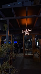 A dimly lit entrance to a sports bar with a glowing neon sign reading 'Good Vibes Only' above the door. The interior appears to be busy, with people visible in the background and various decorations inside. Potted plants are arranged near the entrance, adding a touch of greenery. The wooden door and support beams contribute to a rustic ambiance.