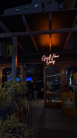 A dimly lit entrance to a sports bar with a glowing neon sign reading 'Good Vibes Only' above the door. The interior appears to be busy, with people visible in the background and various decorations inside. Potted plants are arranged near the entrance, adding a touch of greenery. The wooden door and support beams contribute to a rustic ambiance.