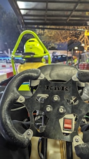 A close-up view from the perspective of a go-kart driver. The steering wheel with a brand logo is prominently displayed in the foreground. Ahead, a person in yellow attire and a bright yellow helmet is seated in another go-kart. The scene is illuminated by overhead lights, and a track with barriers is visible under a metal canopy.