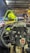 A close-up view from the perspective of a go-kart driver. The steering wheel with a brand logo is prominently displayed in the foreground. Ahead, a person in yellow attire and a bright yellow helmet is seated in another go-kart. The scene is illuminated by overhead lights, and a track with barriers is visible under a metal canopy.