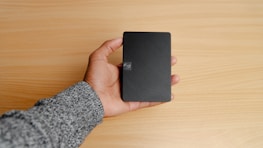A hand holding a small, rectangular black object, possibly a portable external hard drive, against a light wooden surface. The person's sleeve is gray.