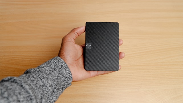 A hand holding a small, rectangular black object, possibly a portable external hard drive, against a light wooden surface. The person's sleeve is gray.