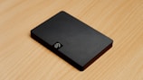 A sleek black external hard drive with a textured diagonal stripe pattern sits on a light wooden surface. The device has a compact rectangular shape with smooth edges, and its surface is glossy, reflecting some light. A small logo is visible on one corner of the hard drive.