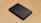 A sleek black external hard drive with a textured diagonal stripe pattern sits on a light wooden surface. The device has a compact rectangular shape with smooth edges, and its surface is glossy, reflecting some light. A small logo is visible on one corner of the hard drive.