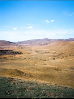 A vast open landscape with rolling hills extending into the distance under a bright blue sky. Sparse vegetation and winding paths are visible, adding texture to the terrain. The scene conveys a sense of spaciousness and tranquility.