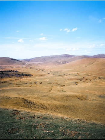 A vast open landscape with rolling hills extending into the distance under a bright blue sky. Sparse vegetation and winding paths are visible, adding texture to the terrain. The scene conveys a sense of spaciousness and tranquility.