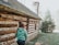 A person wearing a green hoodie walks with a dog near a rustic log cabin featuring stone and wood construction. A chimney is visible on the roof, and a coniferous tree stands in the yard.