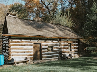 A rustic log cabin with a wooden shingled roof set amidst a background of autumn foliage. The structure is made of horizontal logs, featuring small windows and a simple wooden door. A blue barrel sits outside and a small wooden bench is placed in front of the cabin. The surrounding area has grassy ground with fallen leaves scattered.