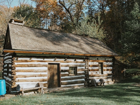 A rustic log cabin with a wooden shingled roof set amidst a background of autumn foliage. The structure is made of horizontal logs, featuring small windows and a simple wooden door. A blue barrel sits outside and a small wooden bench is placed in front of the cabin. The surrounding area has grassy ground with fallen leaves scattered.