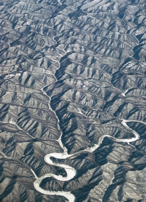 An aerial view of a rugged landscape with numerous ridges and valleys. A winding river snakes through the terrain, creating a stark contrast against the surrounding earth. The textures of the hills and the flowing river suggest a dramatic geological formation.