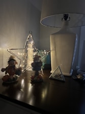 A dimly lit scene on a wooden surface featuring various decorative items. On the left, there are two small scarecrow-like figurines dressed in autumn attire surrounded by small pumpkins. In the center, there is a lighted star-shaped wireframe decoration with fairy lights inside, and behind it, a textured vase or lantern. On the right, there is a large white table lamp with a white lampshade and a small triangular glass terrarium containing seashells.