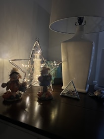 A dimly lit scene on a wooden surface featuring various decorative items. On the left, there are two small scarecrow-like figurines dressed in autumn attire surrounded by small pumpkins. In the center, there is a lighted star-shaped wireframe decoration with fairy lights inside, and behind it, a textured vase or lantern. On the right, there is a large white table lamp with a white lampshade and a small triangular glass terrarium containing seashells.