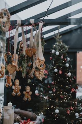 A festive scene with a decorated Christmas tree adorned with lights, red and white ornaments, and a variety of hanging gingerbread cookies and breadsticks. Heart-shaped cookies and pretzels hang from burlap ribbons, and a table displays wrapped gifts and candles. The setting is cozy with a warm, inviting atmosphere.