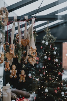 A festive scene with a decorated Christmas tree adorned with lights, red and white ornaments, and a variety of hanging gingerbread cookies and breadsticks. Heart-shaped cookies and pretzels hang from burlap ribbons, and a table displays wrapped gifts and candles. The setting is cozy with a warm, inviting atmosphere.