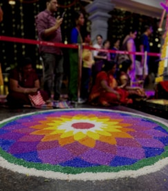 A vibrant and colorful rangoli pattern is displayed on the floor, featuring an intricate design with petals radiating outward. The design is made up of vibrant hues including purple, yellow, orange, and green. In the background, several people are gathered, some standing and others seated, appearing to be engaged in a cultural or festive event. Decorative lights and a red barrier can be seen behind them.