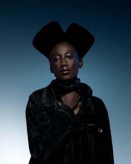 A person with a unique hairstyle stands confidently against a gradient blue background. The individual is wearing a dark jacket and a scarf around the neck, with their hand placed near their collar. The lighting creates a dramatic and polished look.