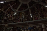 A ceiling with wooden beams and dim lighting houses numerous hanging woven baskets and handicrafts. The shelves display an array of colorful figurines and pottery items. The scene has an eclectic and rustic atmosphere.