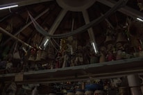 A ceiling with wooden beams and dim lighting houses numerous hanging woven baskets and handicrafts. The shelves display an array of colorful figurines and pottery items. The scene has an eclectic and rustic atmosphere.