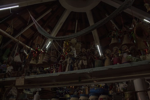 A ceiling with wooden beams and dim lighting houses numerous hanging woven baskets and handicrafts. The shelves display an array of colorful figurines and pottery items. The scene has an eclectic and rustic atmosphere.