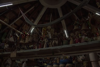 A ceiling with wooden beams and dim lighting houses numerous hanging woven baskets and handicrafts. The shelves display an array of colorful figurines and pottery items. The scene has an eclectic and rustic atmosphere.