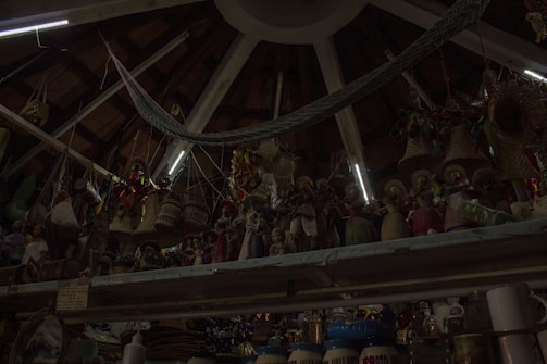 A ceiling with wooden beams and dim lighting houses numerous hanging woven baskets and handicrafts. The shelves display an array of colorful figurines and pottery items. The scene has an eclectic and rustic atmosphere.