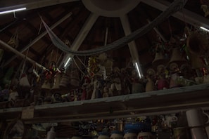 A ceiling with wooden beams and dim lighting houses numerous hanging woven baskets and handicrafts. The shelves display an array of colorful figurines and pottery items. The scene has an eclectic and rustic atmosphere.