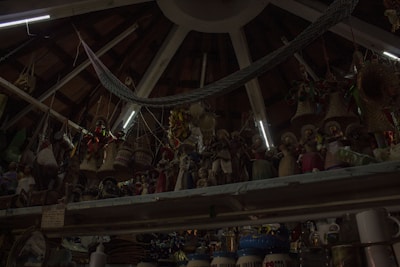 A ceiling with wooden beams and dim lighting houses numerous hanging woven baskets and handicrafts. The shelves display an array of colorful figurines and pottery items. The scene has an eclectic and rustic atmosphere.