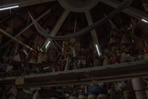 A ceiling with wooden beams and dim lighting houses numerous hanging woven baskets and handicrafts. The shelves display an array of colorful figurines and pottery items. The scene has an eclectic and rustic atmosphere.