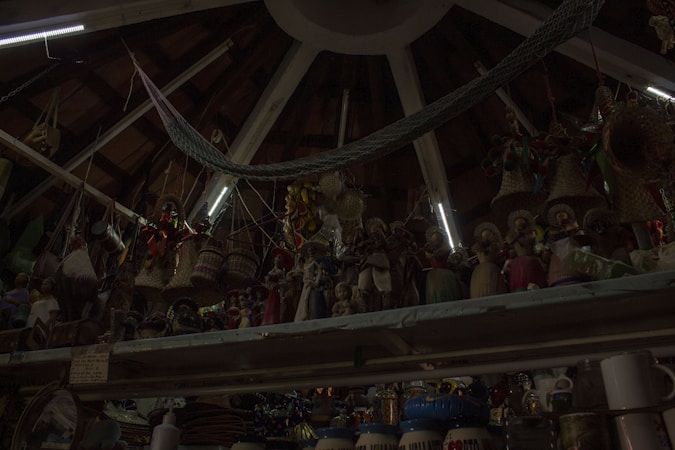 A ceiling with wooden beams and dim lighting houses numerous hanging woven baskets and handicrafts. The shelves display an array of colorful figurines and pottery items. The scene has an eclectic and rustic atmosphere.