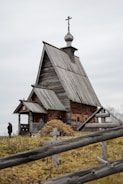 An old wooden church with a steep roof and a cross-topped cupola stands in a grassy area. A wooden fence with simple cross markers surrounds the building. A person dressed in dark clothing is visible near the church.