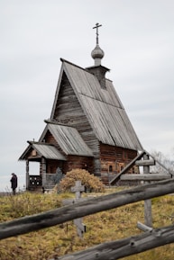 An old wooden church with a steep roof and a cross-topped cupola stands in a grassy area. A wooden fence with simple cross markers surrounds the building. A person dressed in dark clothing is visible near the church.