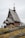An old wooden church with a steep roof and a cross-topped cupola stands in a grassy area. A wooden fence with simple cross markers surrounds the building. A person dressed in dark clothing is visible near the church.