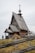 An old wooden church with a steep roof and a cross-topped cupola stands in a grassy area. A wooden fence with simple cross markers surrounds the building. A person dressed in dark clothing is visible near the church.