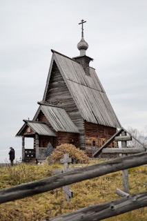 An old wooden church with a steep roof and a cross-topped cupola stands in a grassy area. A wooden fence with simple cross markers surrounds the building. A person dressed in dark clothing is visible near the church.