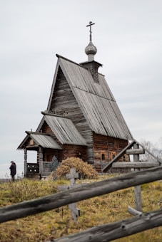 An old wooden church with a steep roof and a cross-topped cupola stands in a grassy area. A wooden fence with simple cross markers surrounds the building. A person dressed in dark clothing is visible near the church.